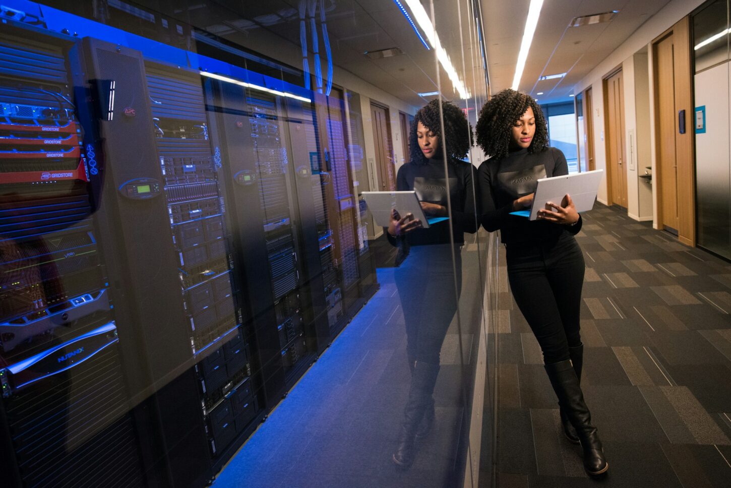 A photo of a woman standing next to a server room and holding a laptop